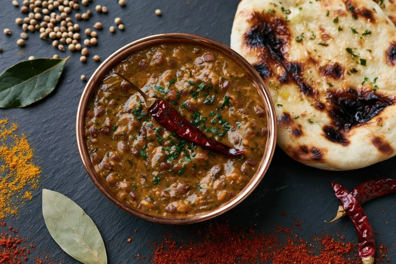 Copper bowl of dal makhani with naan bread and spices arranged on dark slate surface