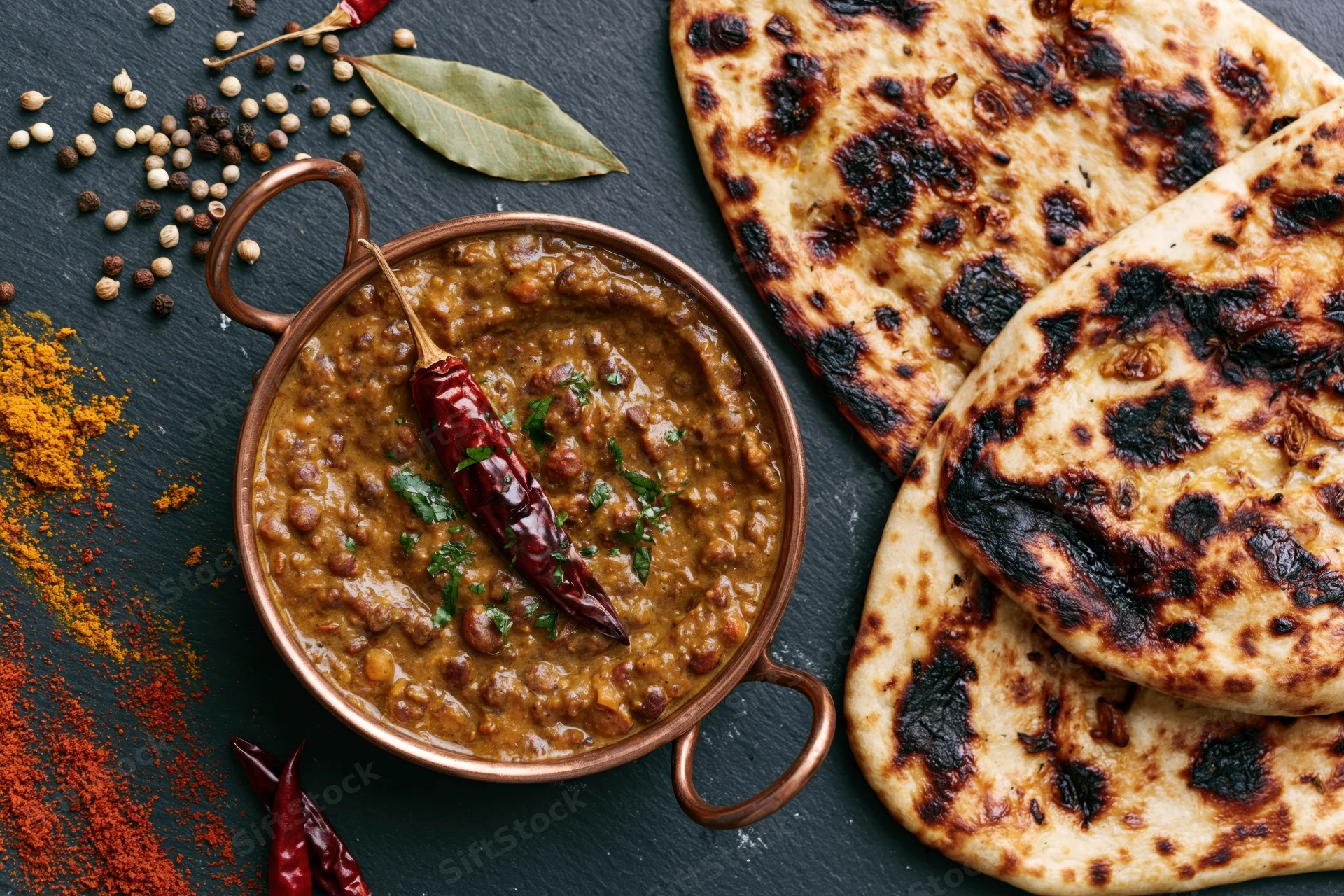 Dal makhani curry in a copper bowl with naan bread and spices arranged on a dark slate surface