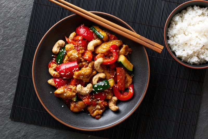 Stir fry with chicken, cashews, vegetables and sesame seeds in a dark matte ceramic bowl with chopsticks, served with rice on a bamboo placemat