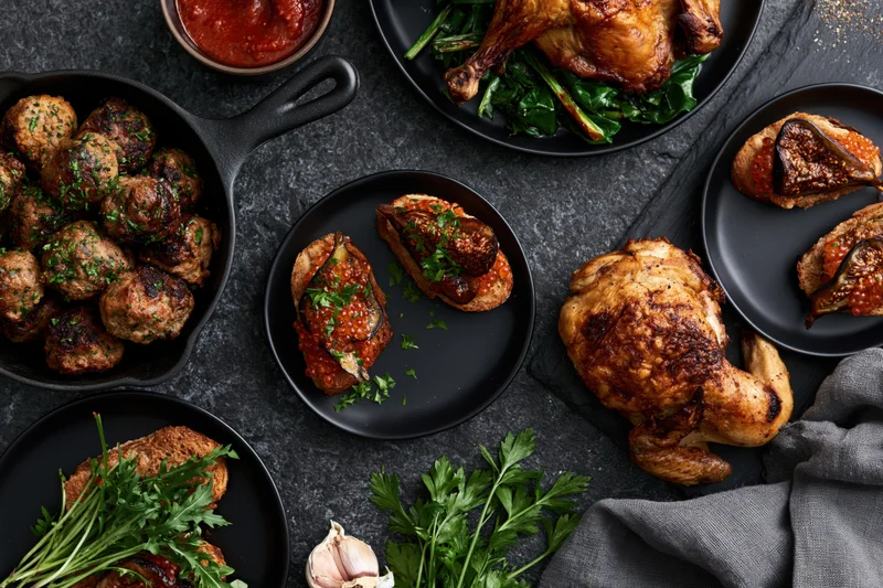 Overhead flat lay of a dinner spread with roasted chicken, meatballs, fig appetizers and herbs on dark slate surface
