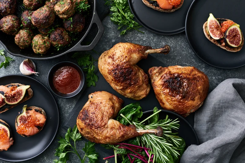 Roasted chicken pieces and meatballs with fig and salmon appetizers arranged on dark slate surface