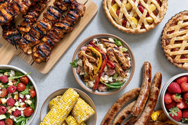 Overhead flat lay of grilled chicken skewers, sausages, corn, salads, pies and strawberries on a gray countertop