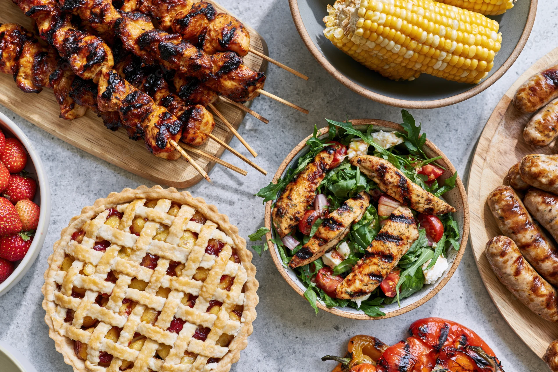 Grilled chicken skewers, corn, sausages, and lattice pie arranged on a gray table with salad and strawberries for a summer barbecue spread