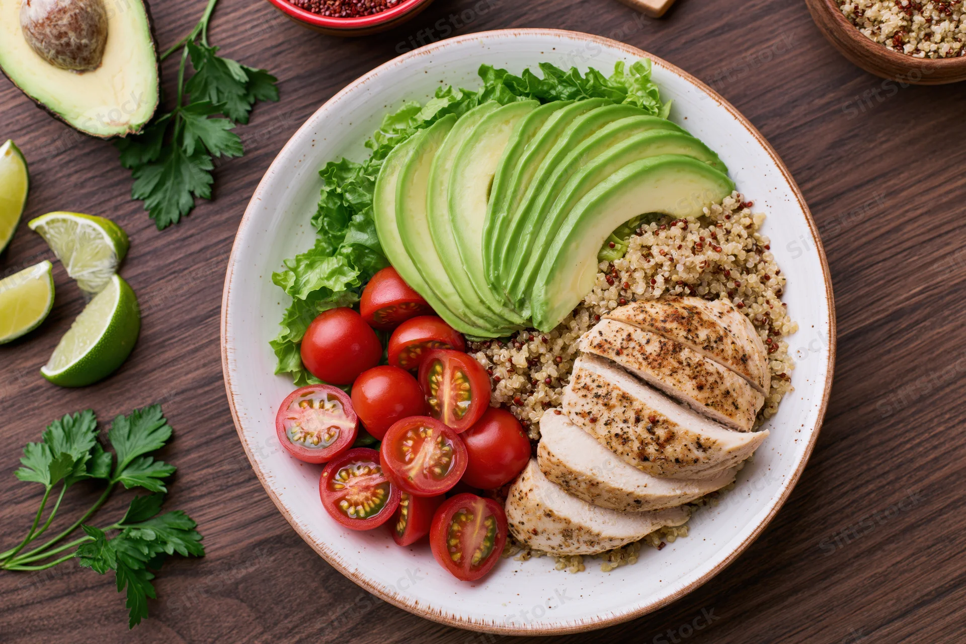 Overhead view of a buddha bowl with quinoa, sliced avocado, grilled chicken, cherry tomatoes, and lettuce on a wooden table
