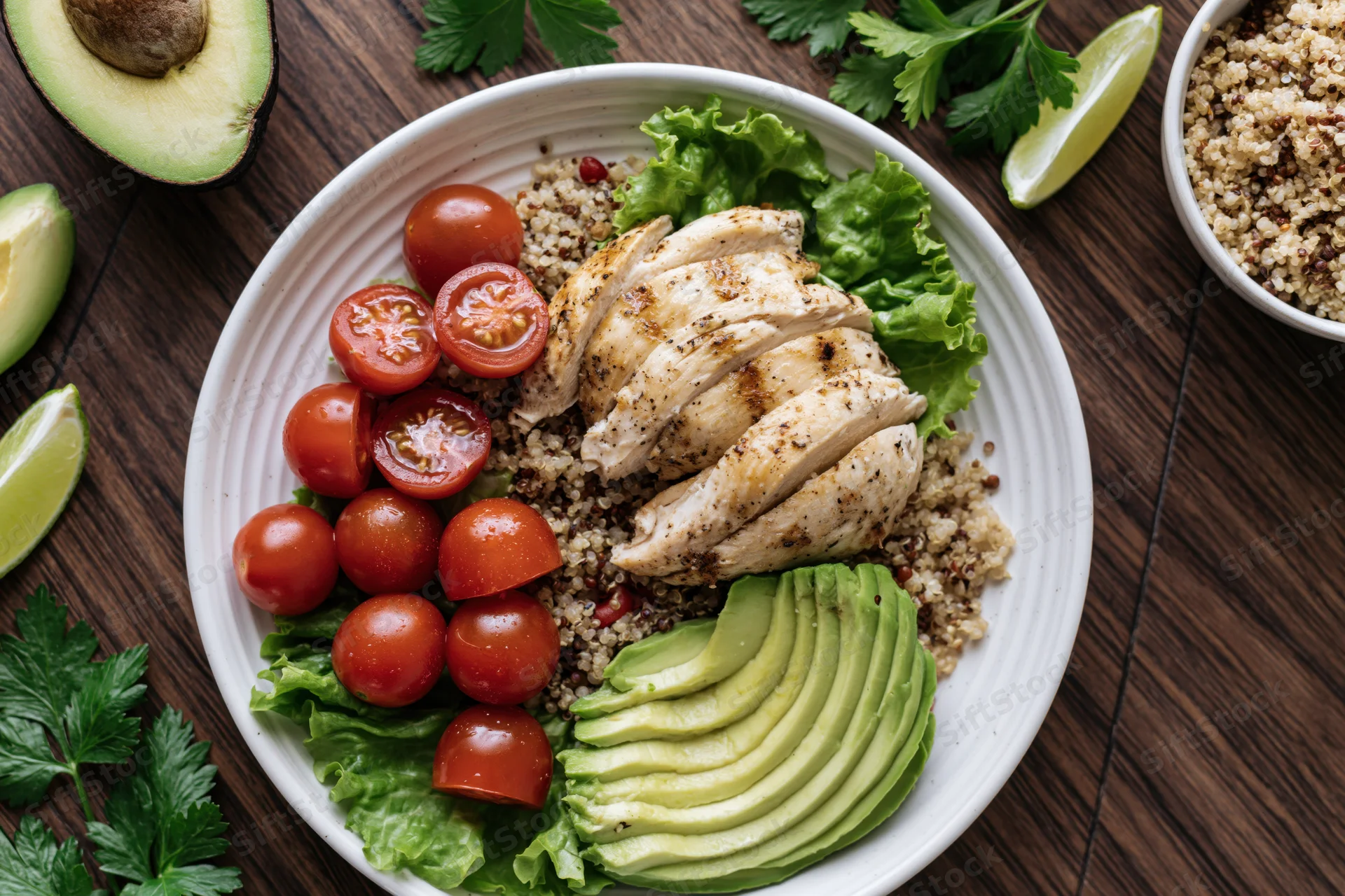 Overhead view of a buddha bowl with quinoa, grilled chicken, avocado, cherry tomatoes, and lettuce on a wooden table