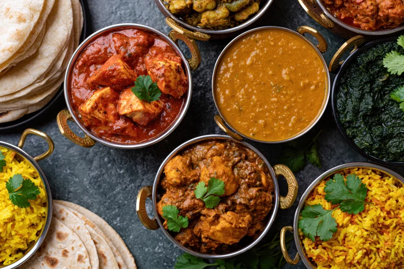 Overhead view of an indian feast with multiple curries, rice, naan and garnishes arranged on a dark surface