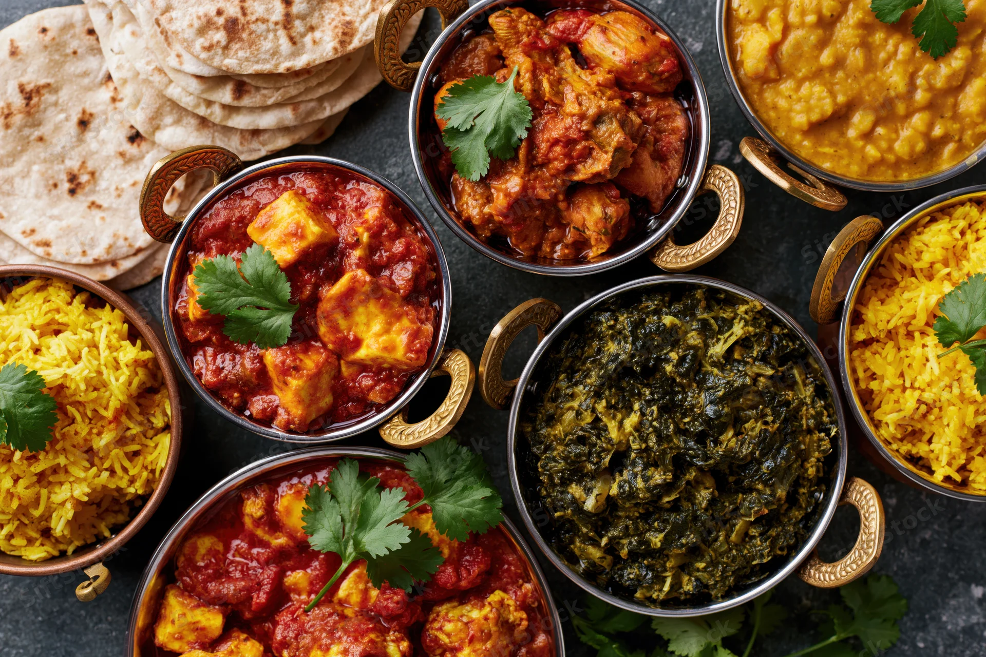 Overhead view of an indian feast spread with curry dishes, rice, naan, and cilantro garnish on a dark stone surface