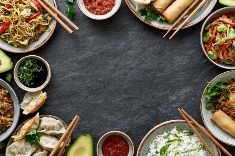 Assorted asian dishes arranged in a circular flat lay on dark slate surface with central copyspace