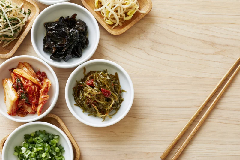 Assorted korean banchan side dishes in white bowls and wooden trays on a light wood table with chopsticks