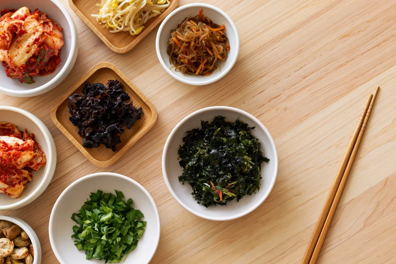 Overhead flat lay of assorted korean banchan side dishes arranged on wooden table with chopsticks