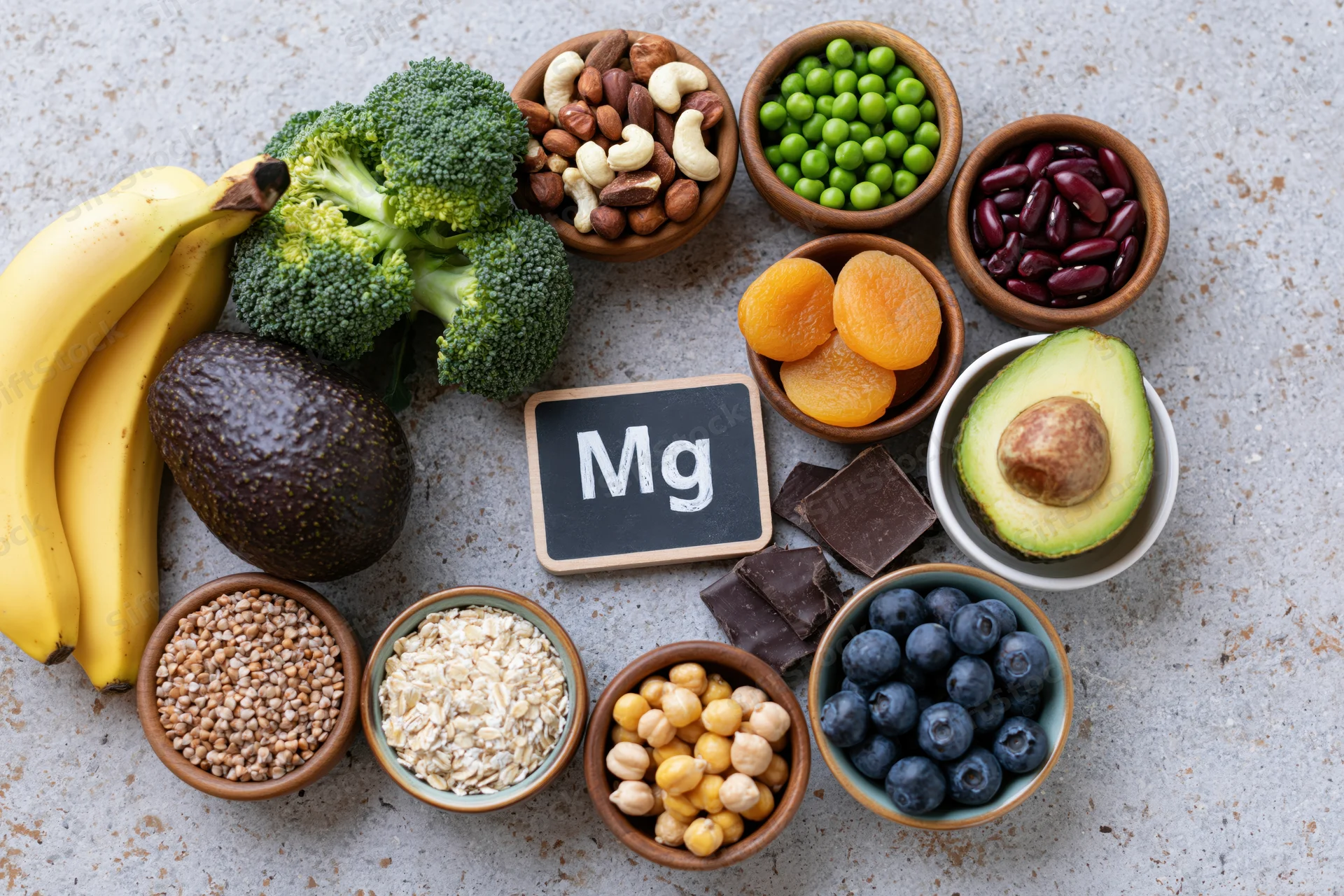 Overhead flat lay of magnesium-rich foods arranged on a gray surface with bowls of nuts, seeds, fruits, vegetables, and dark chocolate surrounding a chalkboard sign labeled Mg