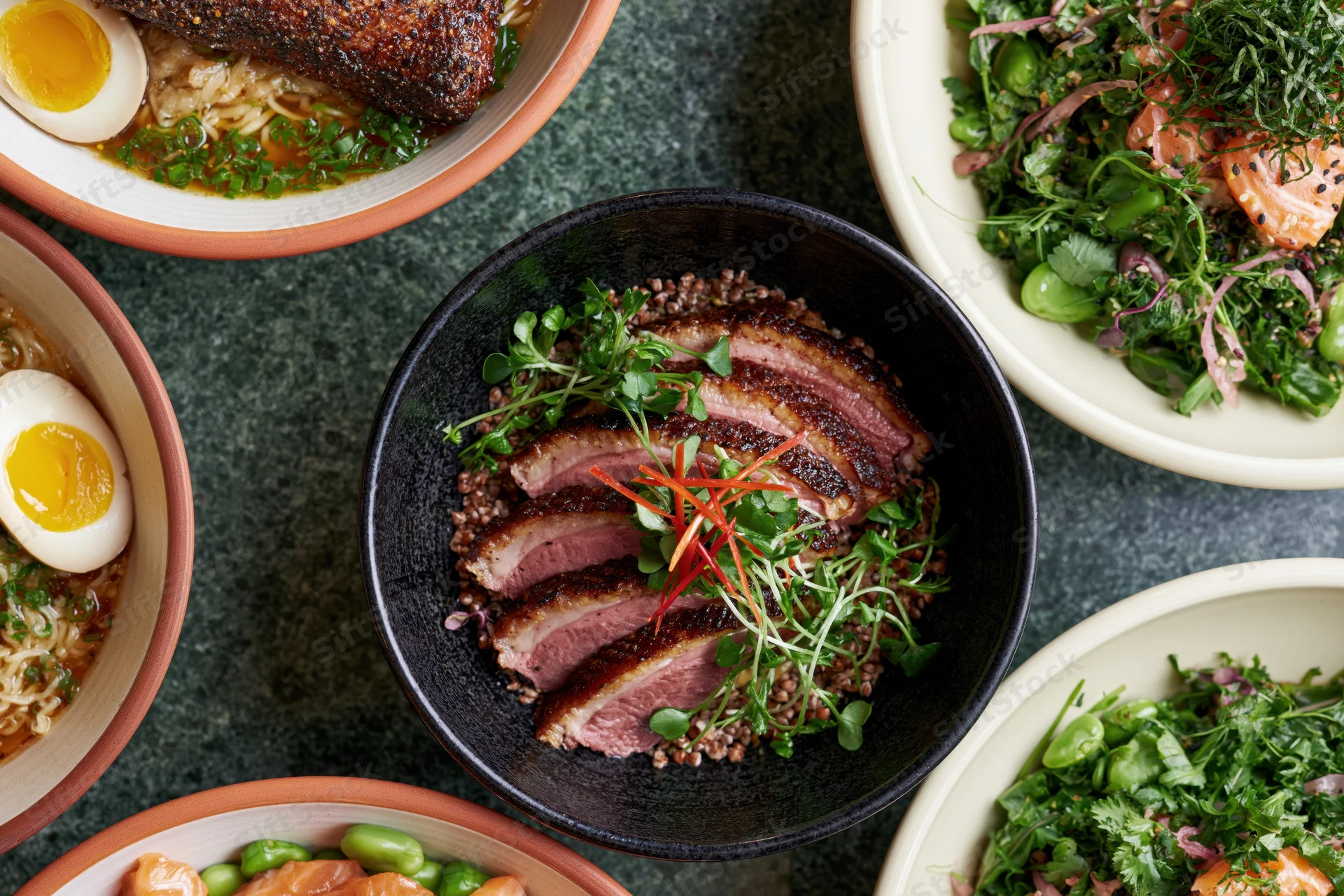 Overhead flat lay of six asian fusion dishes arranged on dark surface with bowls of ramen, salad, and duck on grains
