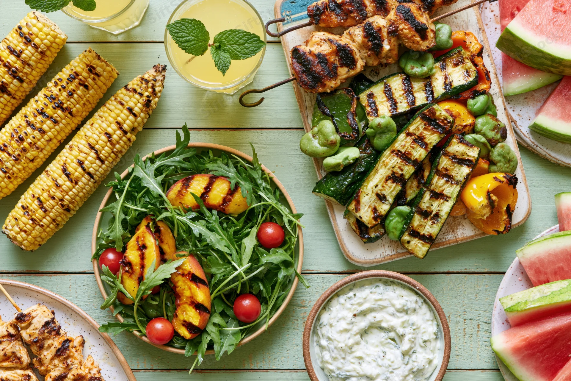 Overhead flat lay of grilled summer barbecue spread on pale green wooden table with corn, vegetables, skewers, salad, watermelon, lemonade and dip