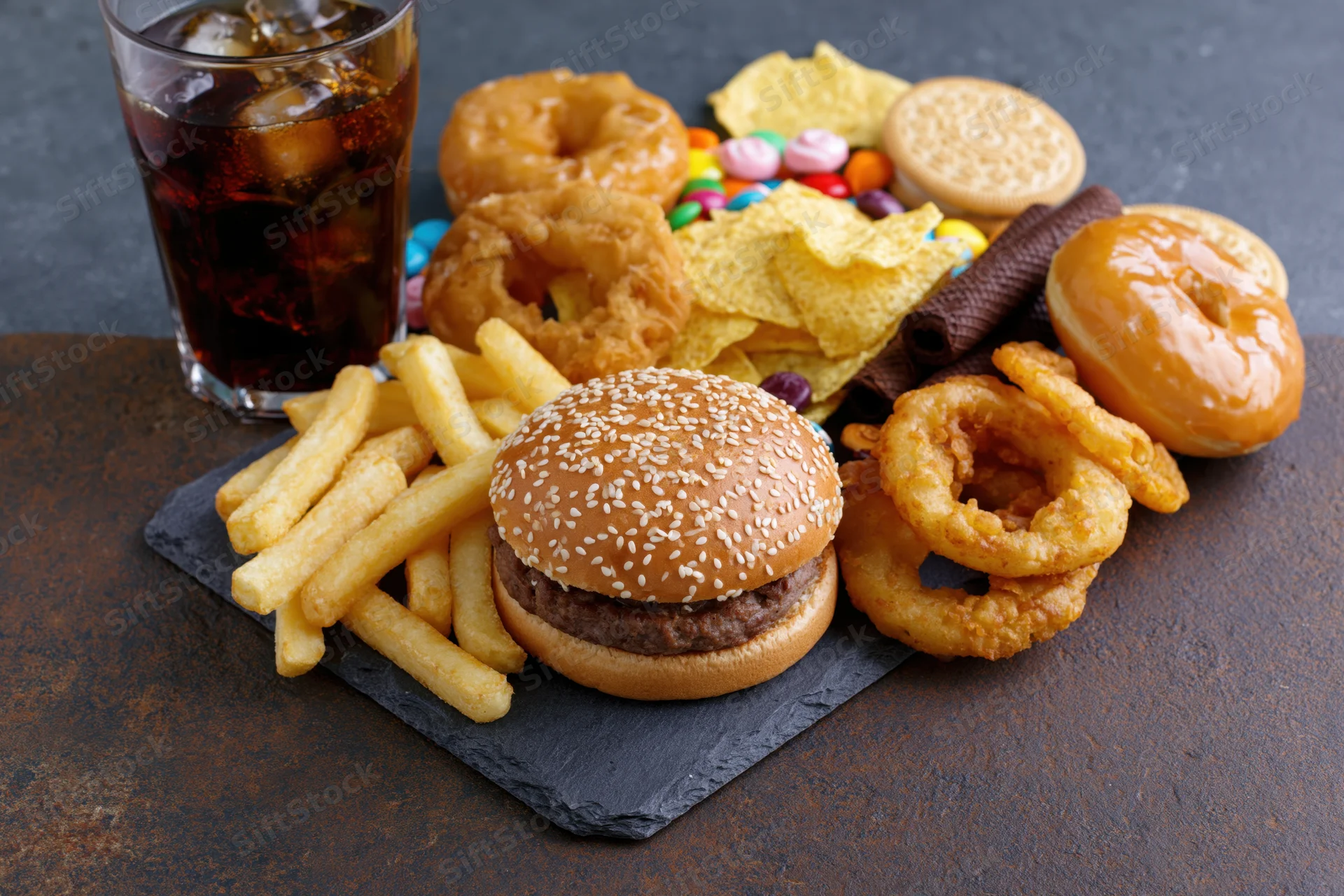 Overhead spread of junk food including burger, fries, soda, onion rings, donuts, chips and candy arranged on dark slate board