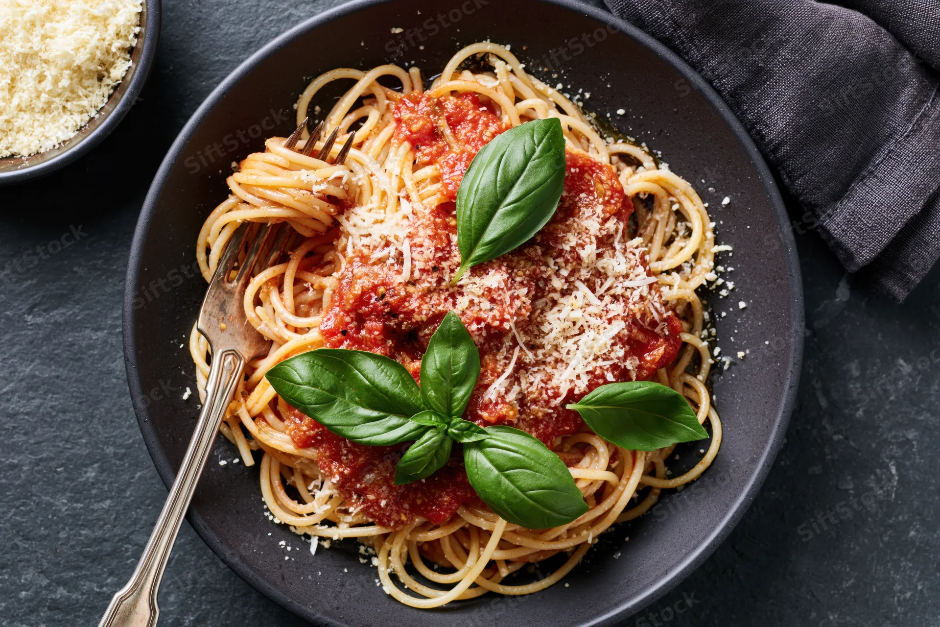 Spaghetti with tomato sauce and grated parmesan in a black bowl, garnished with fresh basil leaves, served with a fork on a dark surface