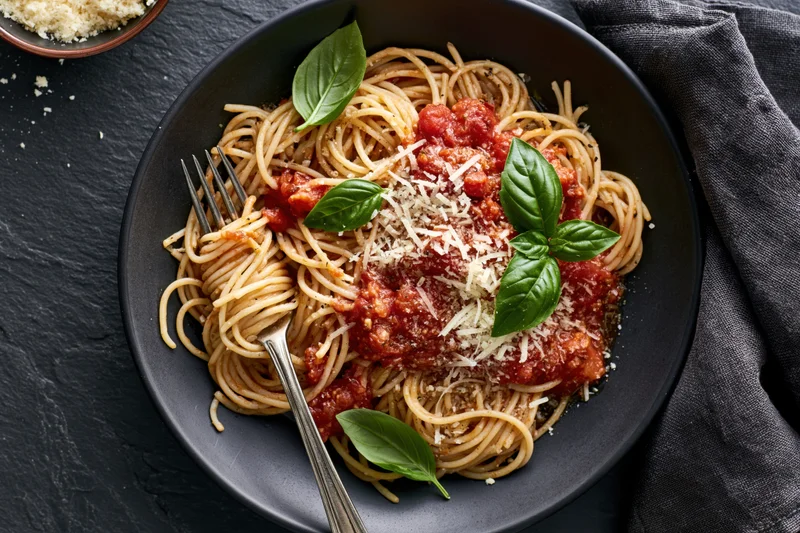 Spaghetti with tomato sauce and grated parmesan on a black plate, garnished with basil leaves, served with a fork on a dark surface