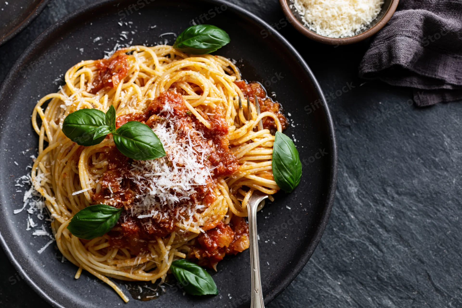 Spaghetti with tomato sauce and grated parmesan on a black plate, garnished with basil leaves, served with a fork on a dark stone surface