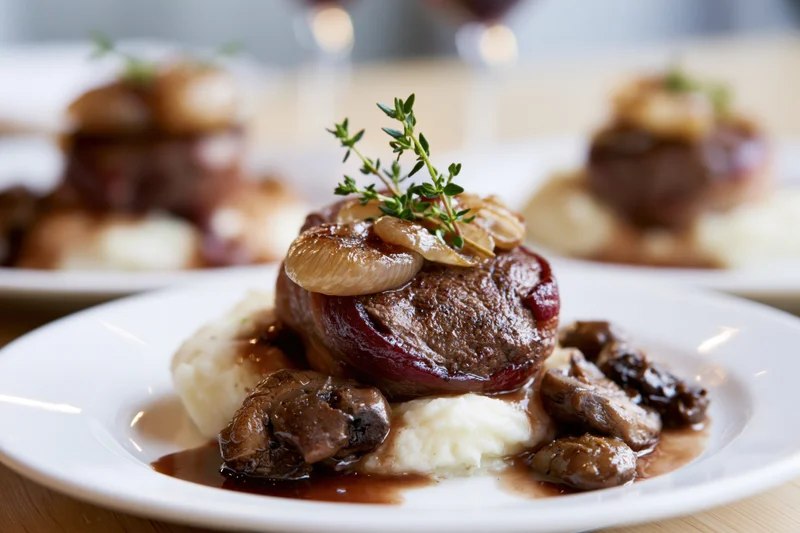Beef medallion wrapped in bacon served on mashed potatoes with mushrooms and gravy on a white plate