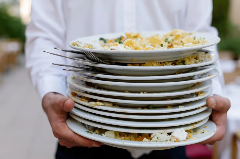 Server holding a tall stack of used white dinner plates with food remnants and cutlery outdoors