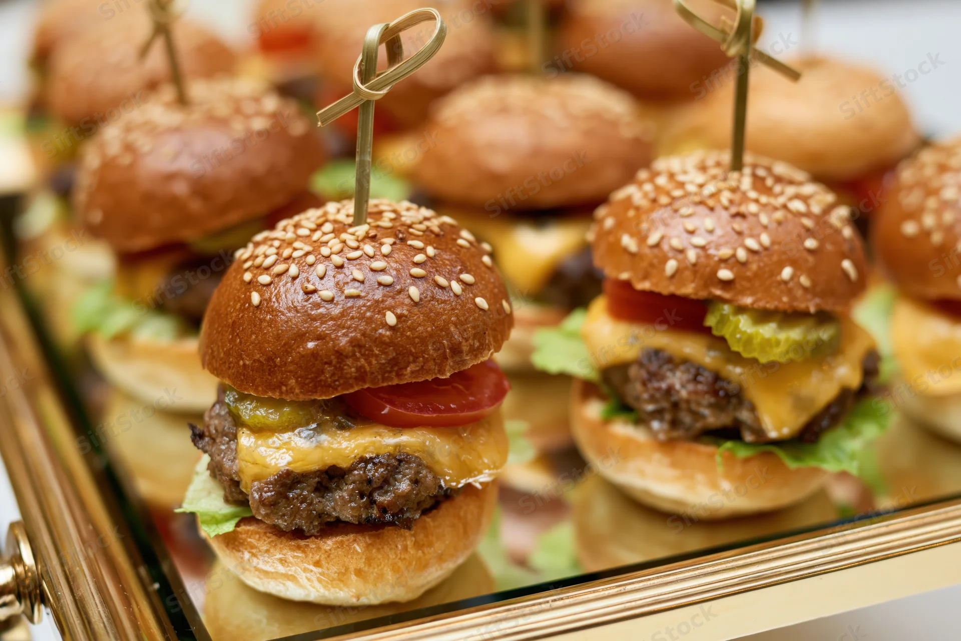 Mini beef sliders with cheese, tomato, lettuce and pickle on sesame buns arranged on a mirrored catering tray