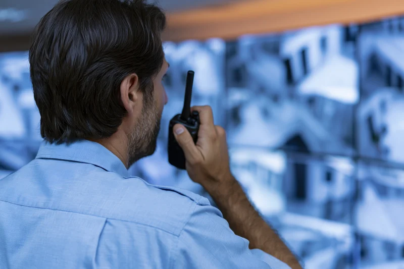 Security guard speaking into handheld radio while monitoring video feeds on a screen wall