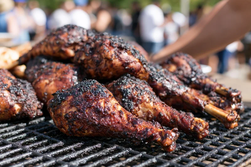 Grilled turkey legs piled on a barbecue grill outdoors with people in the background