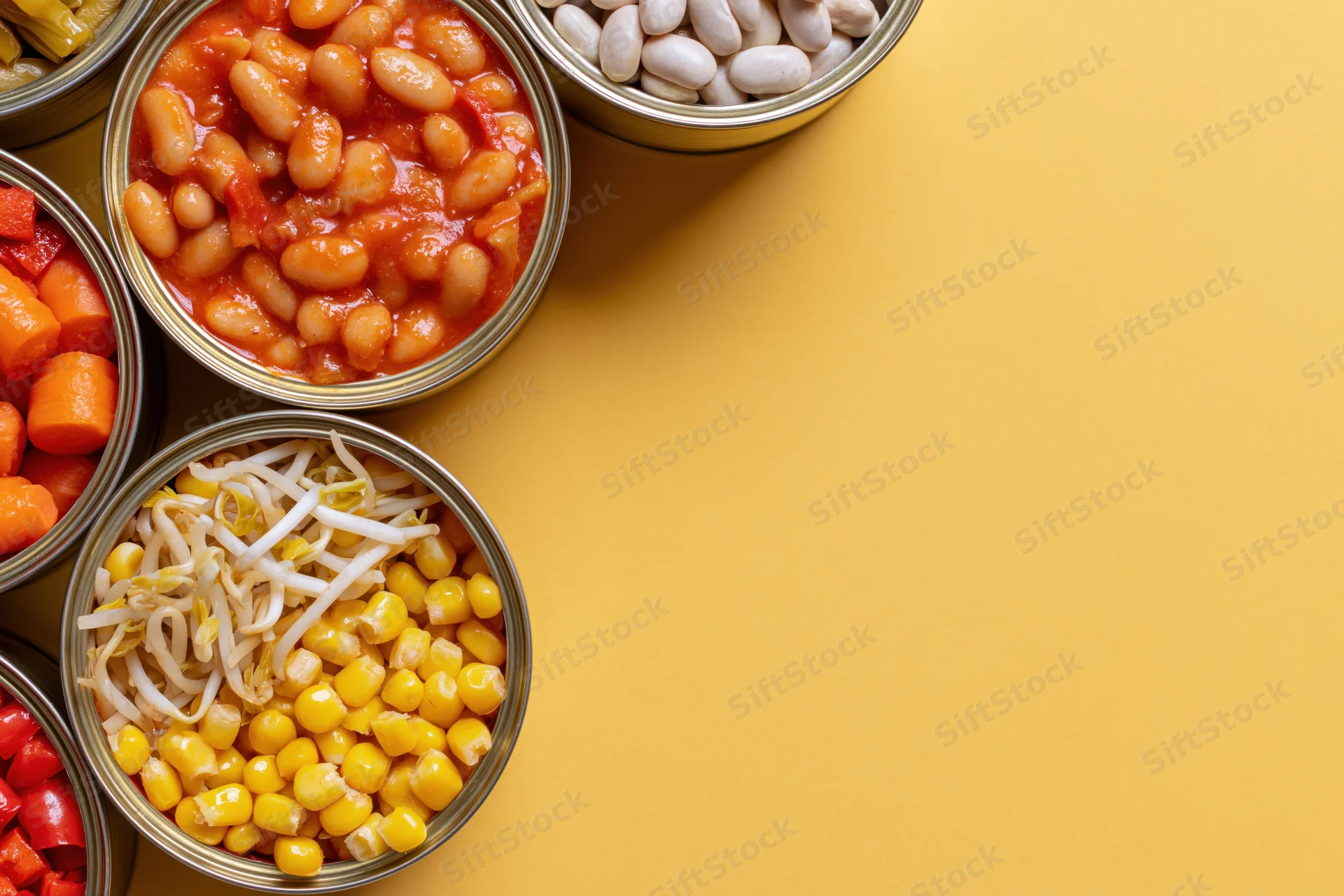 Open tin cans filled with various preserved vegetables arranged on a yellow surface