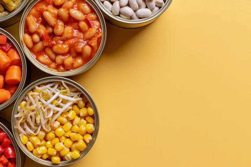 Open tin cans filled with various preserved vegetables arranged on a yellow surface