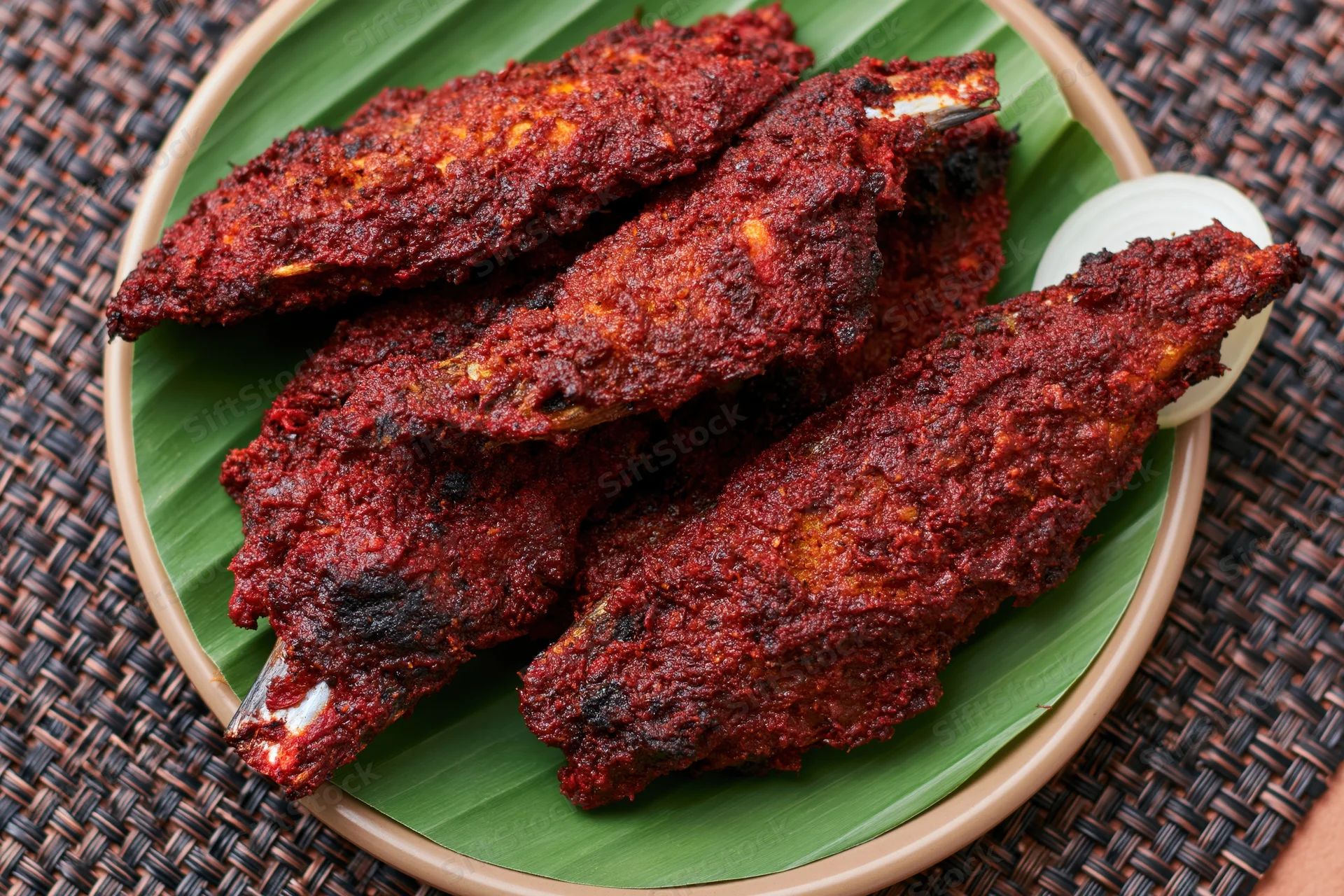 Deep-fried spiced fish coated in dark red curry sauce served on banana leaf in bamboo plate on woven placemat
