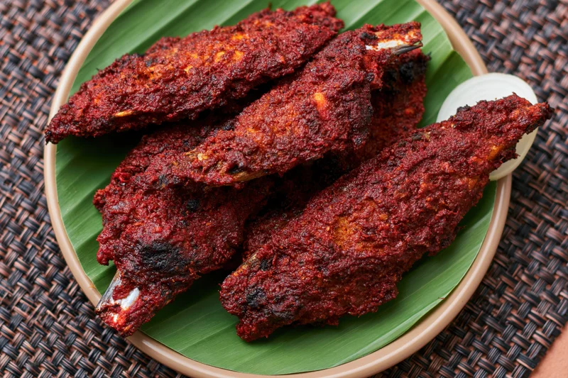 Deep-fried spiced fish coated in dark red curry sauce served on banana leaf in bamboo plate on woven placemat