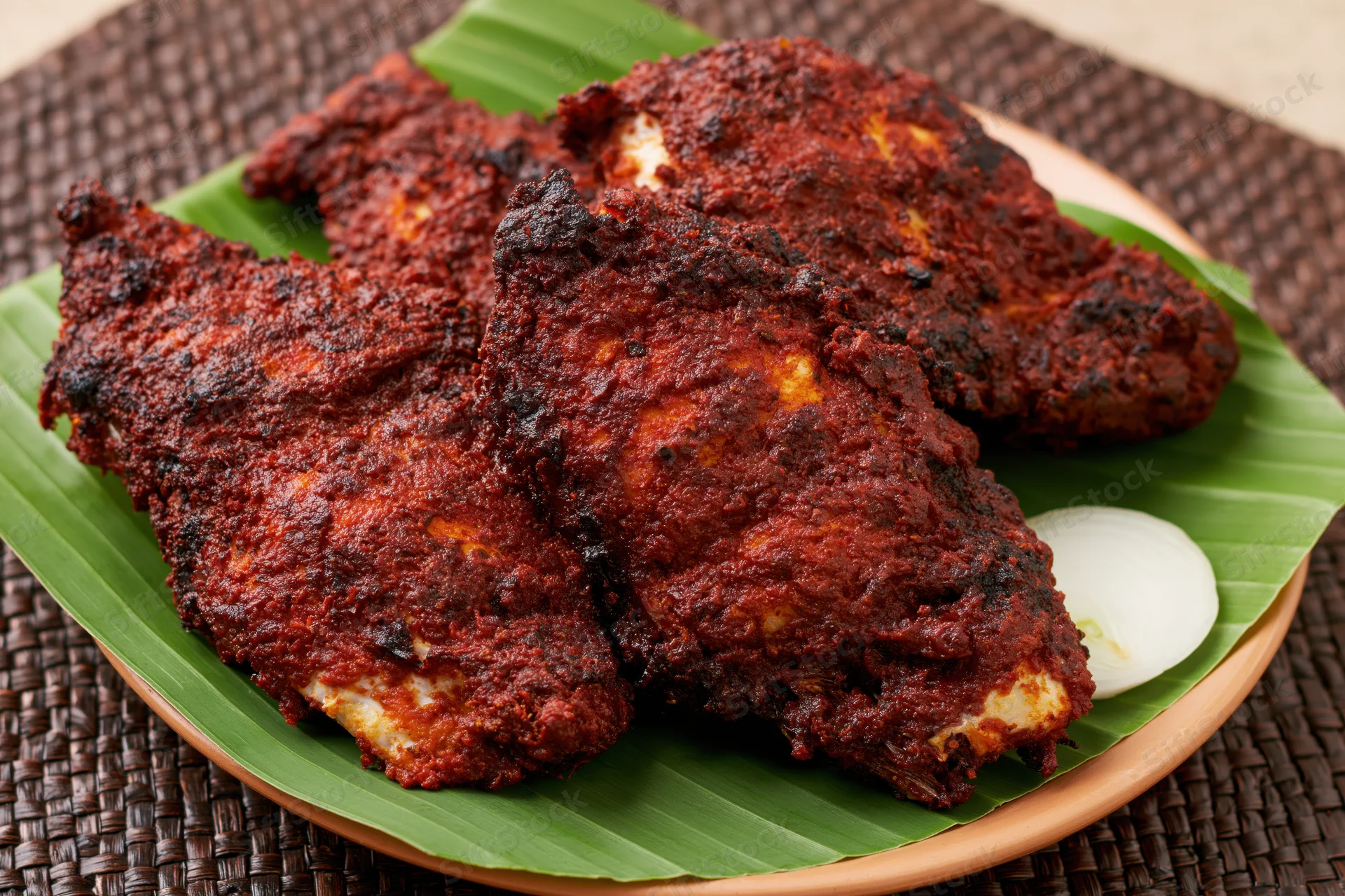 Deep-fried spiced fish pieces served on a banana leaf-lined plate with onion slice on woven mat