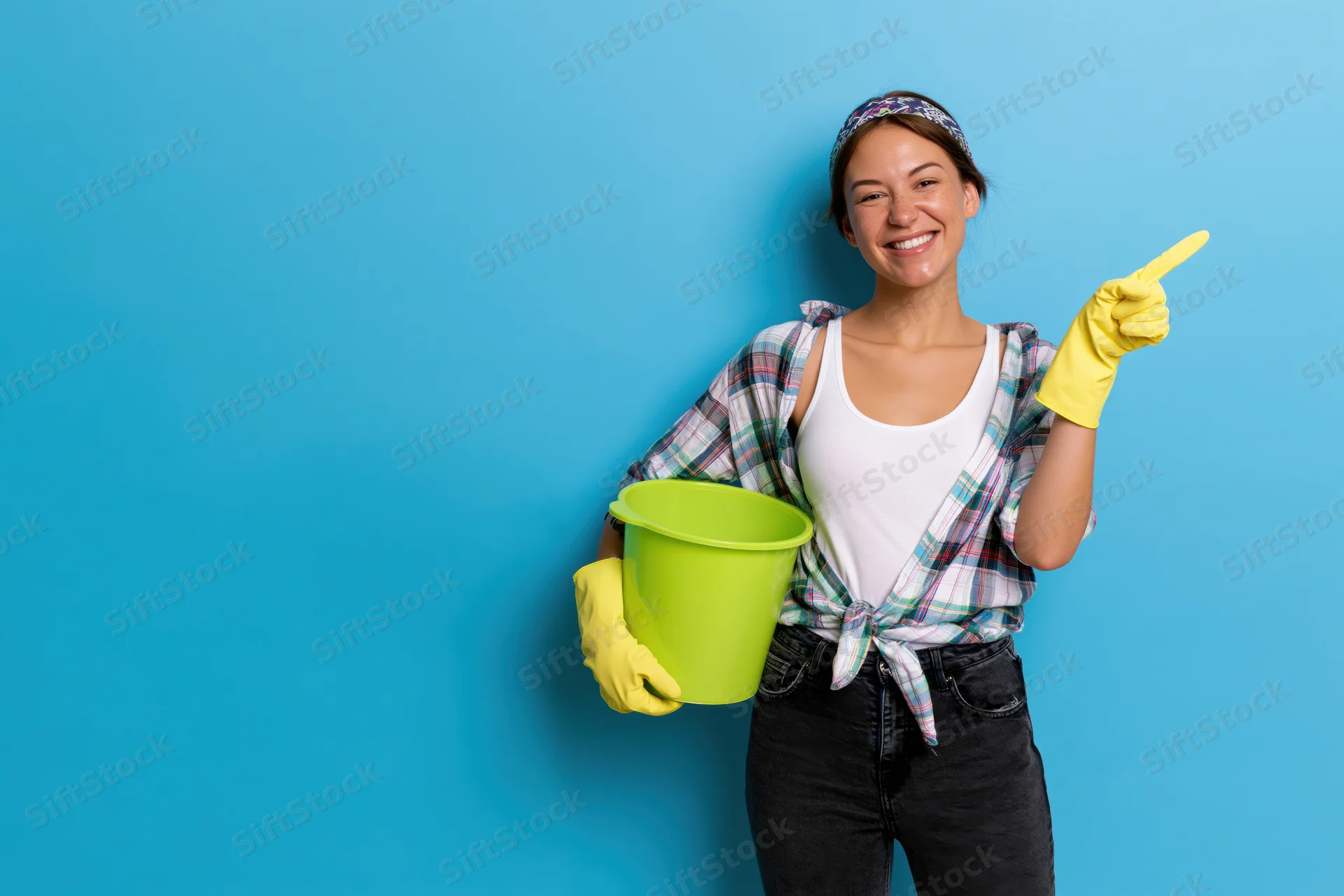 Smiling woman holding green bucket and pointing with yellow glove against blue background