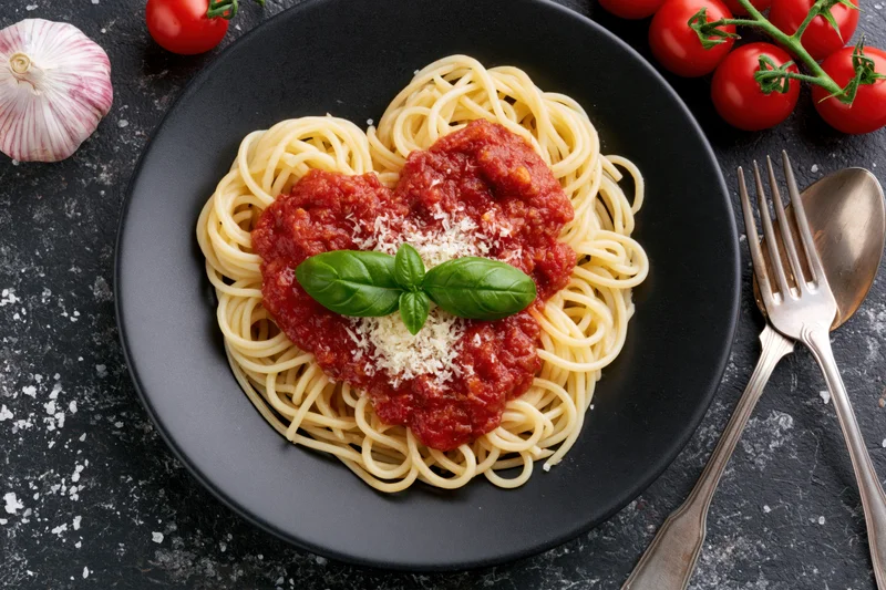 Spaghetti with tomato sauce arranged in heart shape on black plate with basil and parmesan, surrounded by garlic and cherry tomatoes, fork and spoon on dark surface