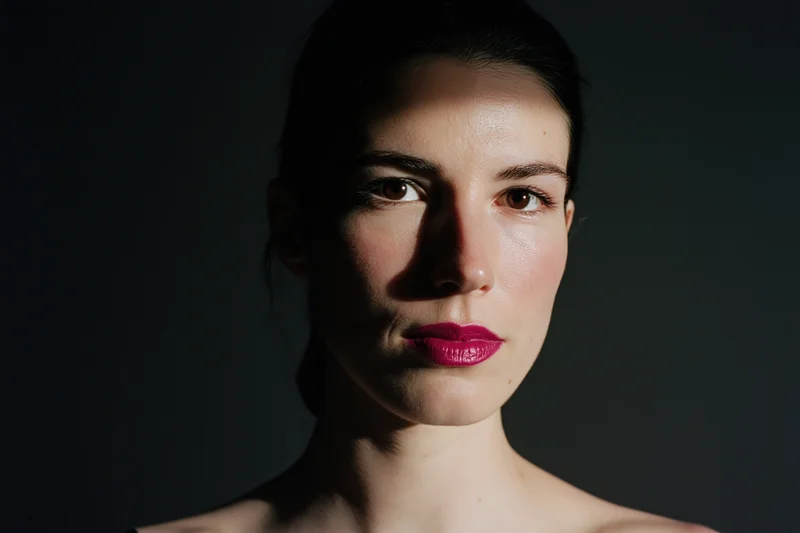 Woman with dark hair slicked back, wearing red lipstick, in dramatic studio lighting