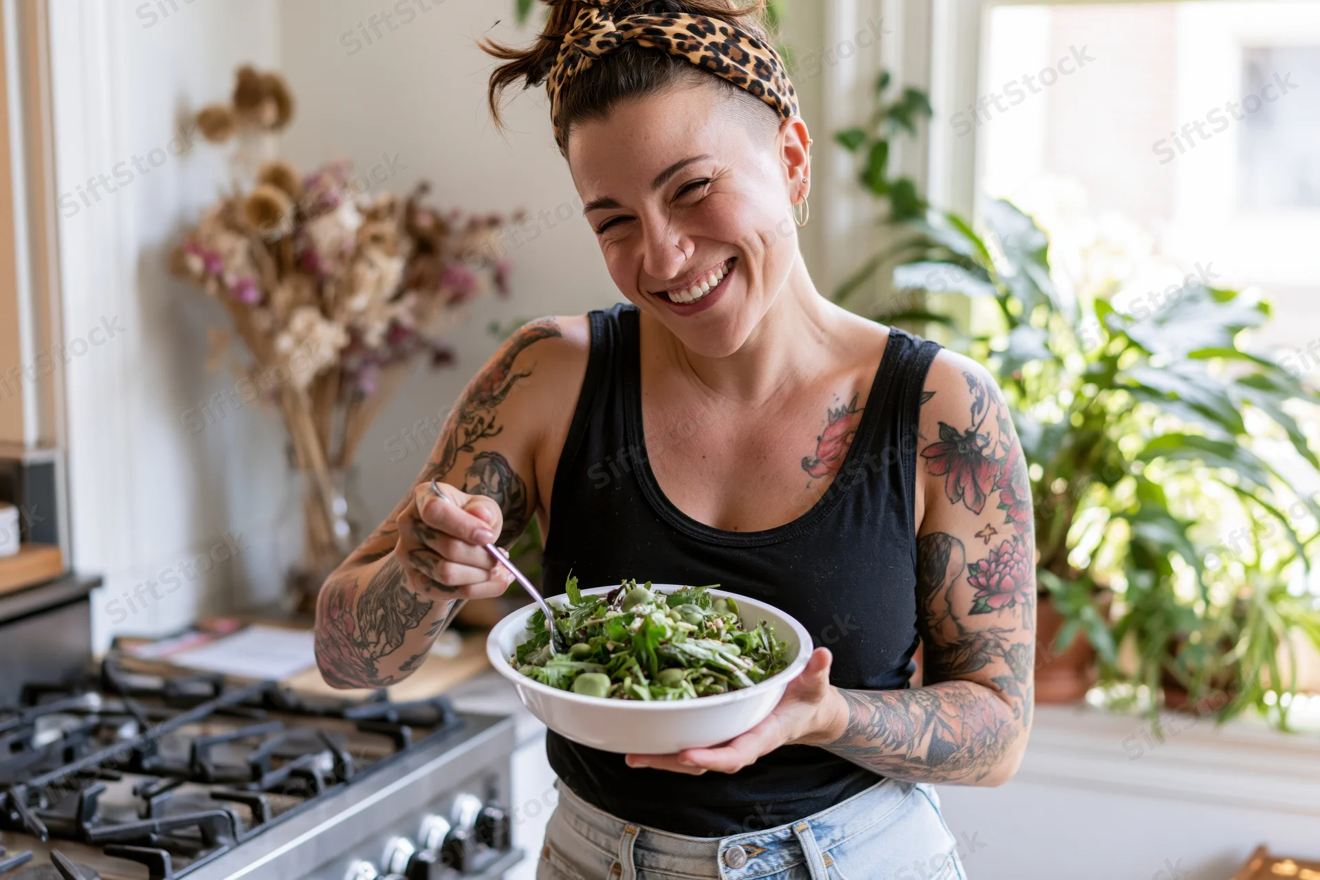 Tattooed woman laughing while holding a bowl of salad in a bright kitchen