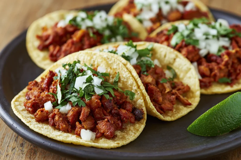 Al pastor tacos on corn tortillas topped with chopped onions and cilantro on a black plate with a lime wedge