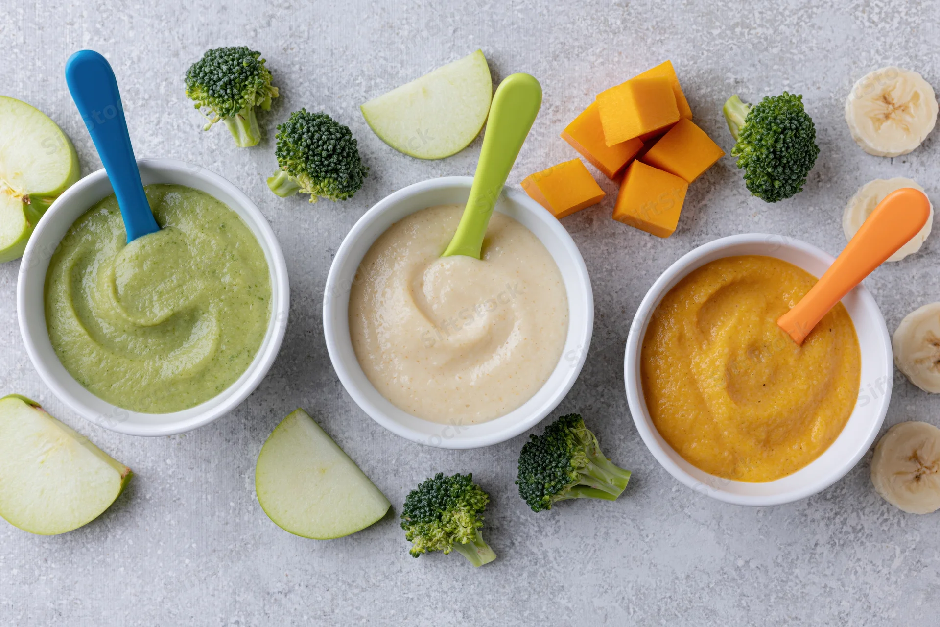 Three white ceramic bowls of baby food puree in green, beige and orange with broccoli, apple, banana and squash arranged on a light surface