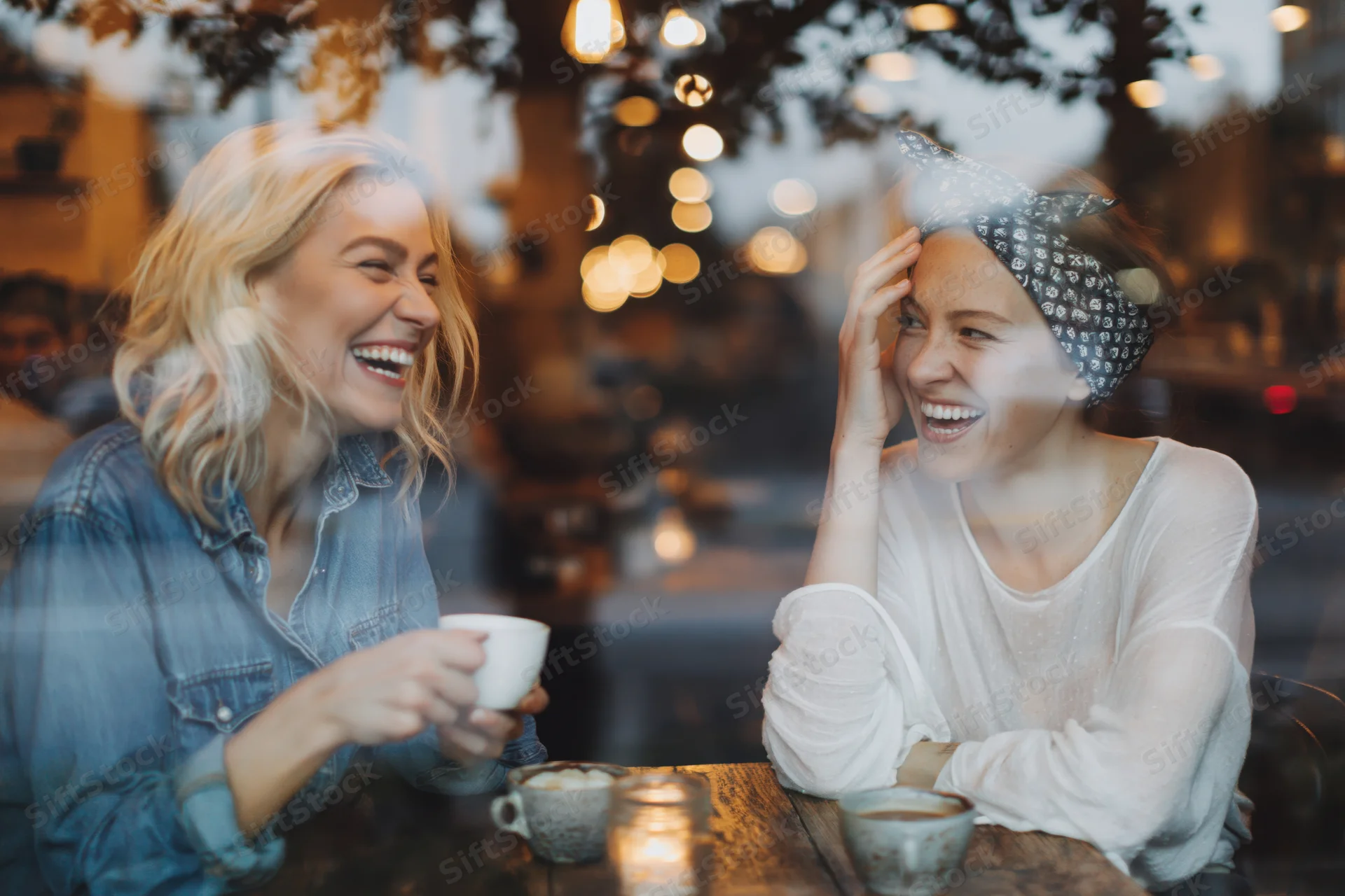 Two women laughing together at a wooden cafe table seen through window reflection