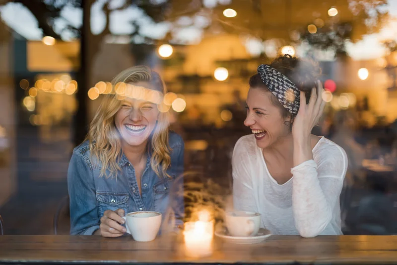 Two women laughing together at a wooden cafe table viewed through glass with warm lights reflecting in the background