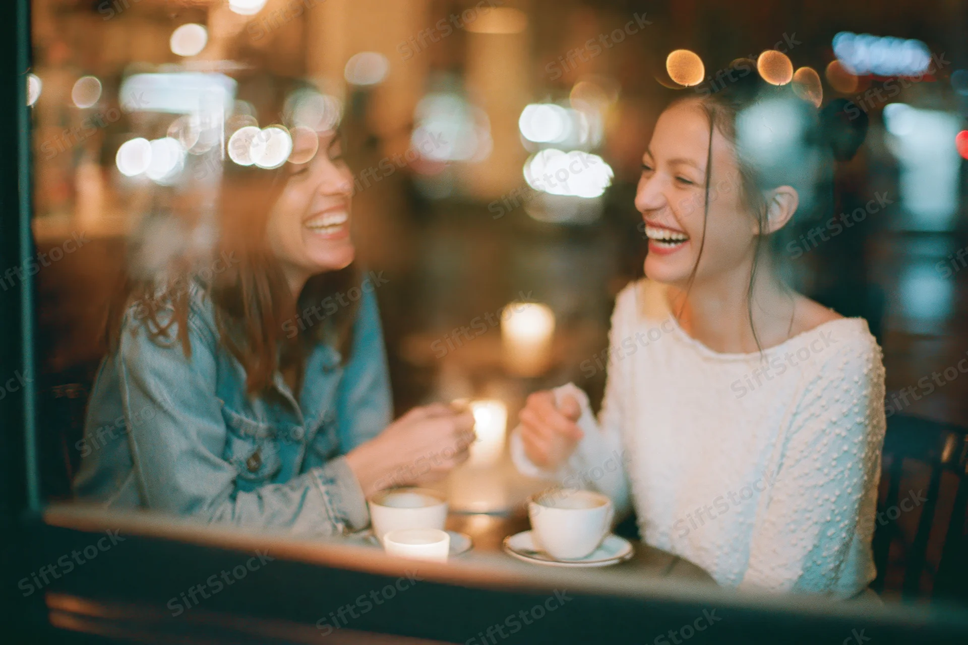 Two women laughing and talking over coffee at a cafe table viewed through a window