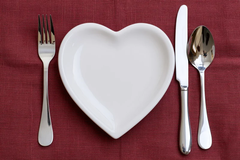 Heart-shaped white ceramic plate centered on red linen tablecloth with silverware arranged around it