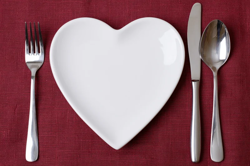 Heart-shaped white ceramic plate centered on red linen table with fork, knife and spoon arranged around it