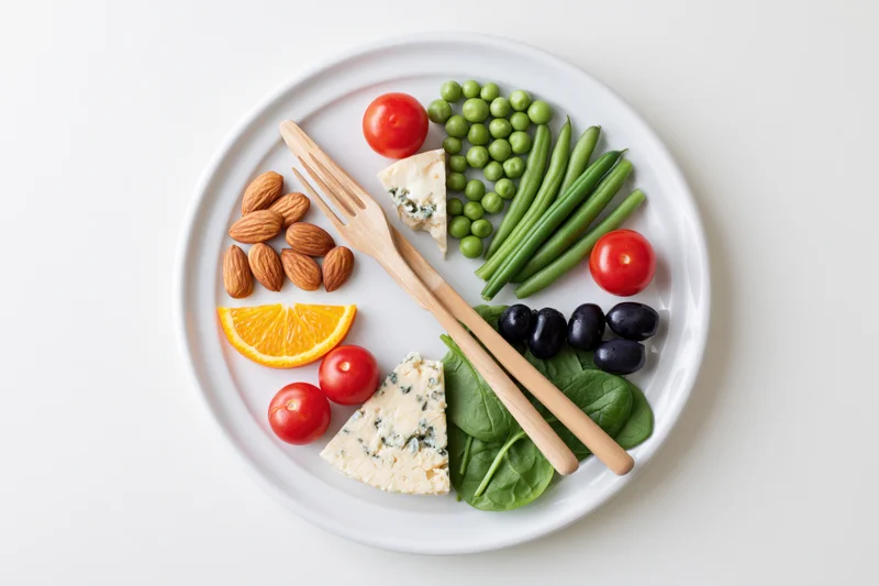White plate with arranged vegetables, fruits, nuts and cheese, viewed from above on a white surface