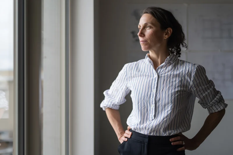 Woman architect standing by window with hands on hips looking thoughtful in office