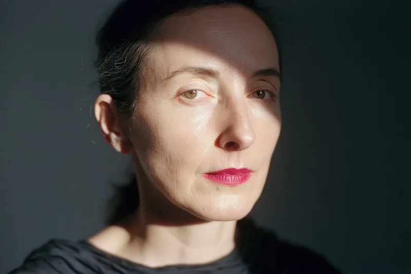 Woman with dark hair pulled back and bright pink lips in soft studio lighting, facing slightly to the side