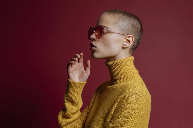 Woman in yellow turtleneck sweater wearing round rose-tinted sunglasses in profile against red background