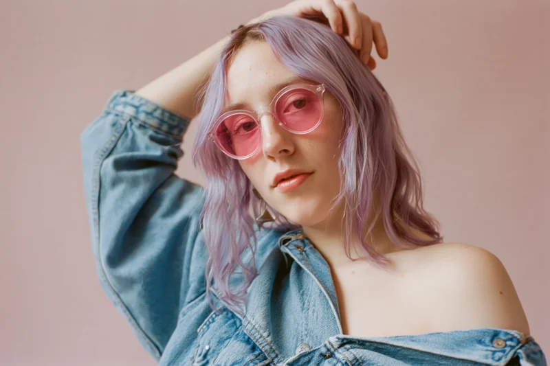 Woman with wavy lavender hair and pink-tinted sunglasses posing in denim jacket against pink background