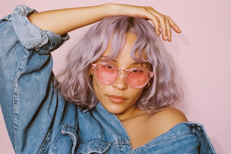Woman with wavy lavender hair and pink-tinted sunglasses wearing denim jacket in studio setting