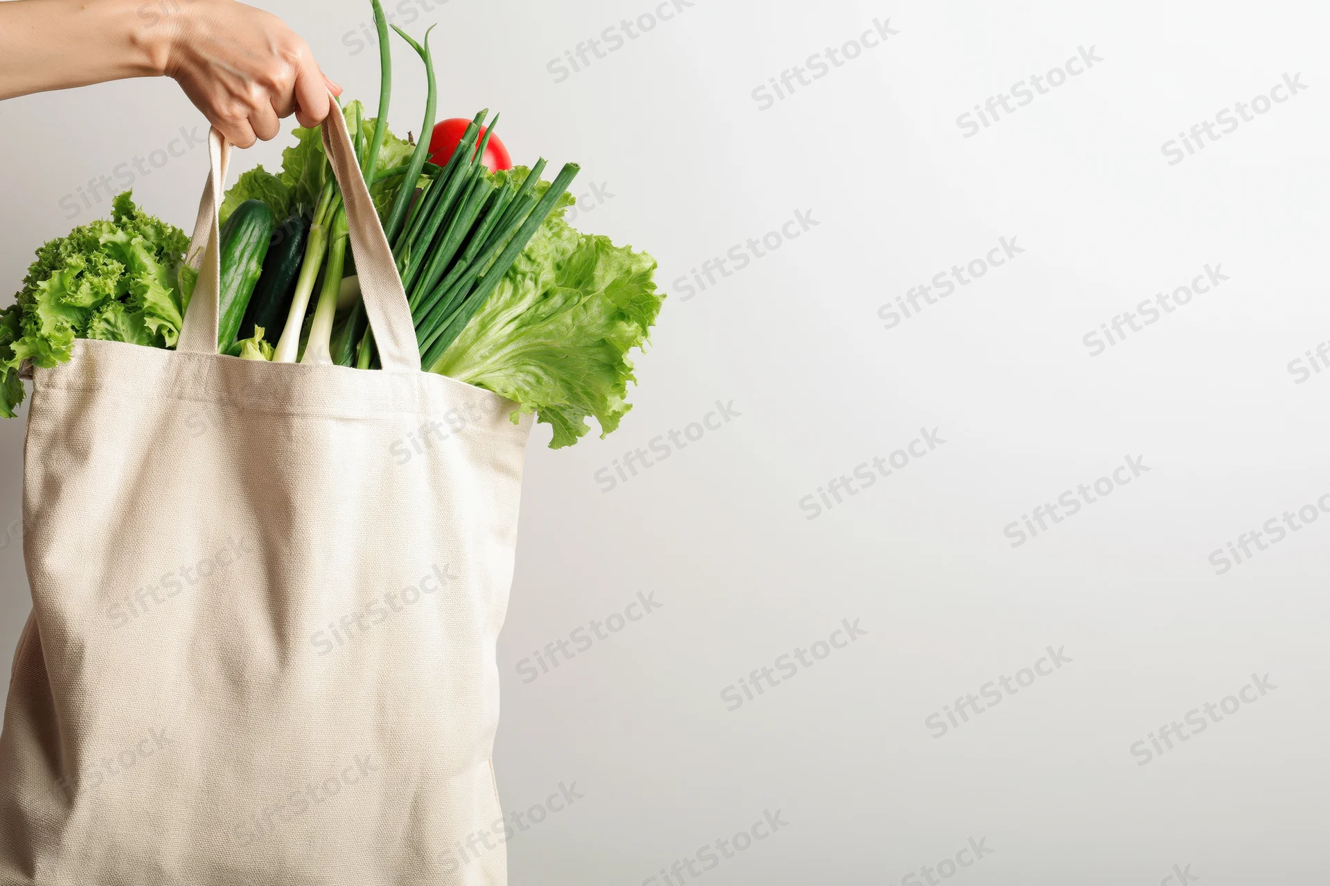 Woman holding a beige cotton tote bag filled with fresh vegetables against a plain background