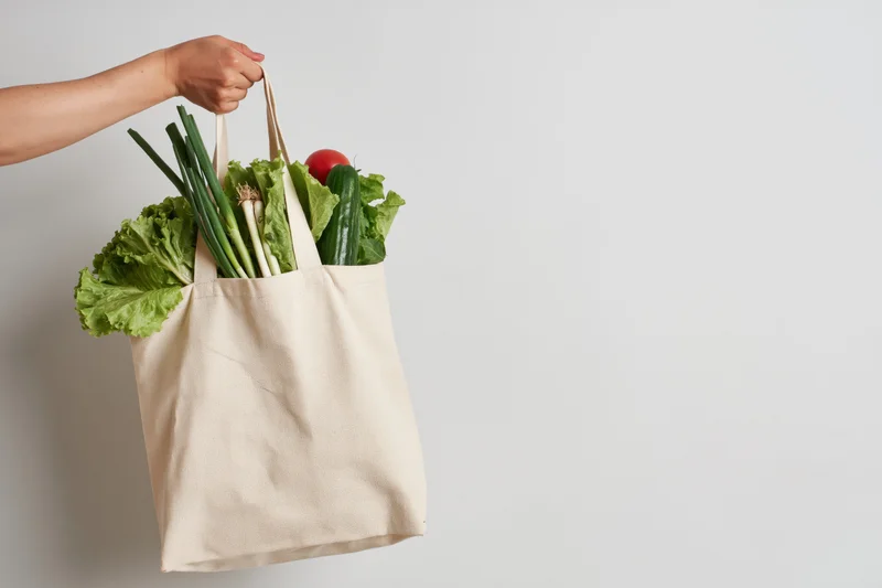 Woman's hand holding a beige cotton tote bag filled with fresh vegetables against a plain background
